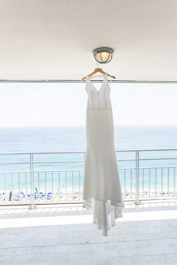 A getting-ready detail shot of a white, minimalist wedding dress hanging from a ceiling light fixture on a wooden hanger, positioned in front of a high-rise balcony with a metal railing. The gown features a deep V-neckline, fitted silhouette, and a slight flare at the hem with a subtle train. The balcony overlooks a busy beach with colorful umbrellas and the ocean visible in the background. The composition is a wide vertical shot, centered on the dress with the beachfront serving as a bright, airy backdrop.