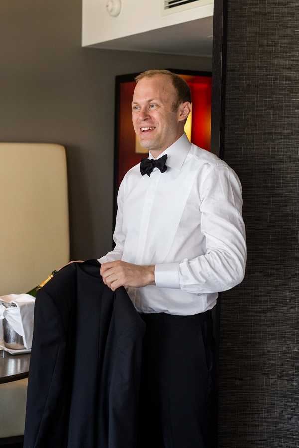 A groom getting ready indoors, holding his dark navy or black tuxedo jacket over his arm while smiling off-camera. He is dressed in a white formal dress shirt with a black bow tie and dark trousers, styled for a classic black-tie wedding. The setting appears to be a modern hotel room with dark textured wall panels and neutral furnishings visible in the background. This is a mid-length portrait shot with soft natural and artificial lighting.