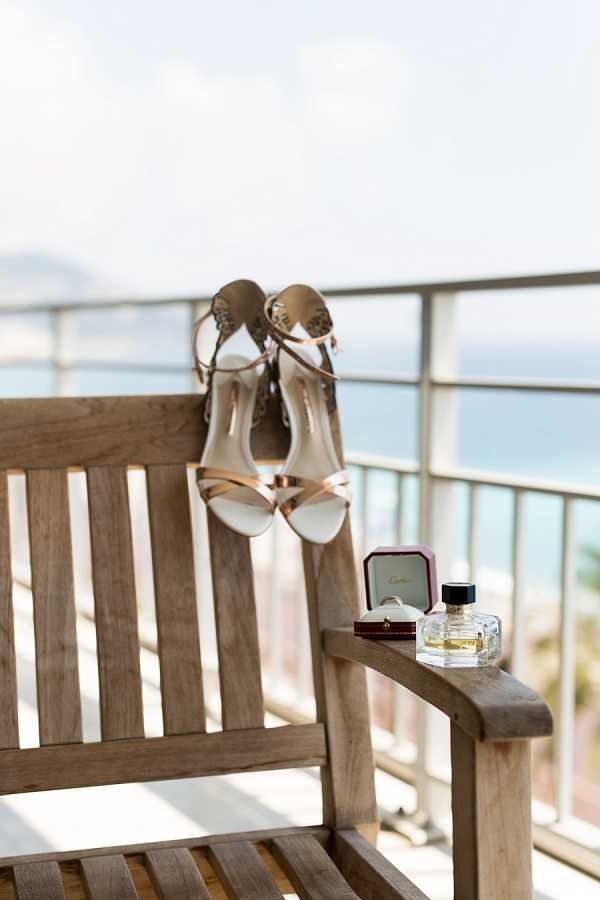 A bridal detail flat lay styled on a wooden outdoor bench on a terrace or balcony overlooking the sea. A pair of gold and ivory strappy heeled sandals with ornate ankle buckles are hooked over the back slat of the bench, while a red Cartier ring box with a gold band visible inside and a small square glass perfume bottle are placed on the armrest. The composition is a mid-range detail shot with the water and a distant coastline softly out of focus in the background.