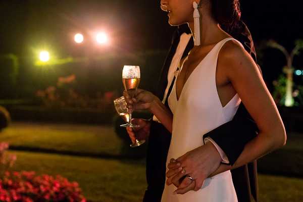 A close-up portrait of a couple outdoors at night during what appears to be a cocktail hour or evening reception. The bride is wearing a sleek white gown with a deep V-neckline and long white tassel earrings, holding a champagne flute with rosé or sparkling wine; she also carries a small black clutch. The groom stands close behind her in a black tuxedo, their hands intertwined, with a wedding band visible on his wrist. Warm artificial uplighting illuminates the scene from behind, set against a manicured garden at night.