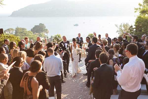 The bride and groom walk back up the aisle together following an outdoor ceremony, with approximately 60 guests lining both sides throwing white flower petals and applauding. The bride wears a fitted, sleeveless white gown and carries a white bouquet, while the groom is dressed in a dark suit. The ceremony took place on a terrace or elevated garden overlooking a large body of water, with a tree-covered peninsula visible in the background. The wide shot captures warm late-afternoon golden light across the scene, with a classic, garden-party styling aesthetic. Potential venue feature image.