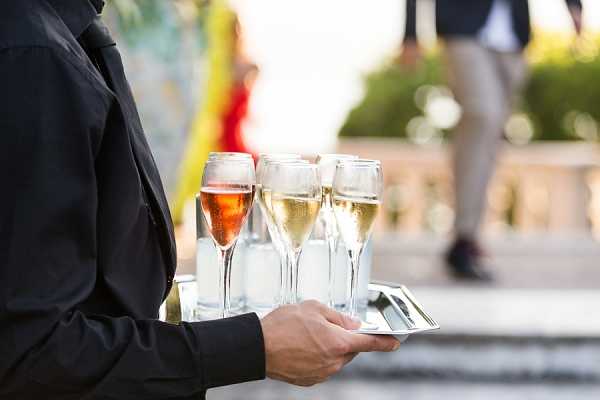 A close-up shot of a server dressed in black holding a polished silver tray with five champagne flutes — one filled with rosé and four with white/brut champagne — during what appears to be an outdoor cocktail hour. The background is softly blurred but shows a stone balustrade terrace with guests visible, including a person in a red outfit and another in beige trousers. The setting appears to be an outdoor terrace, likely at a chateau or formal venue.