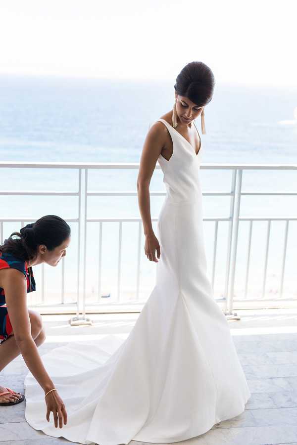A getting-ready moment captured outdoors on a terrace or balcony overlooking the sea, with a white railing and water visible in the background. The bride stands in a minimalist white fitted mermaid-style gown with a deep V-neckline, a structured bodice, and a floor-length train, accessorized with gold tassel earrings and a delicate necklace, her dark hair styled in an updo. A second woman, wearing a red and blue patterned dress, kneels beside her adjusting the train of the gown. The composition is a medium full-length portrait shot with natural daylight, and the overall styling is clean and modern.