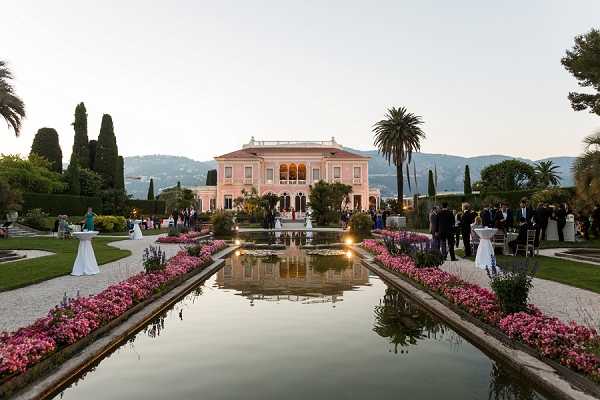 A cocktail hour reception taking place outdoors in the formal gardens of a grand pink Belle Époque villa, shot as a wide landscape image at dusk. A long rectangular reflecting pool runs from the foreground toward the villa, flanked on both sides by densely planted borders of pink and magenta flowering plants and low ground lighting that casts warm reflections on the water. Approximately 40-60 guests in formal attire are mingling near the villa's entrance and terrace, with white cocktail tables dotted across the garden. The pale pink facade of the villa is lit and reflects in the pool, with cypress trees and palm trees framing the architecture against a soft dusk sky. Potential venue feature image.