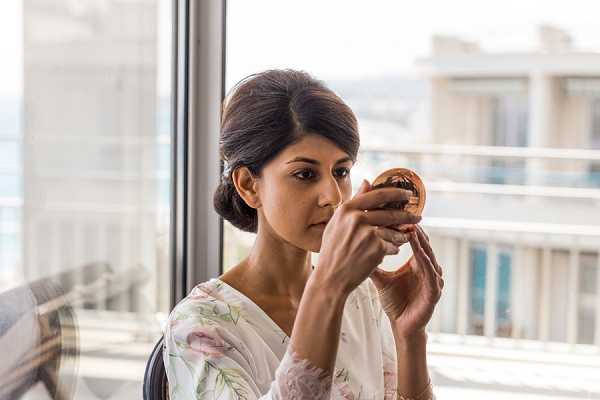 A getting-ready scene showing a bride checking her makeup using a small round hand mirror. She is wearing a white floral robe with soft pink and green botanical print, and her dark hair is styled in a low chignon. The shot is taken indoors in front of large floor-to-ceiling windows with a coastal or urban skyline visible in the background, suggesting a hotel room or high-rise venue. The image is a close-up portrait with natural daylight providing soft, diffused lighting.