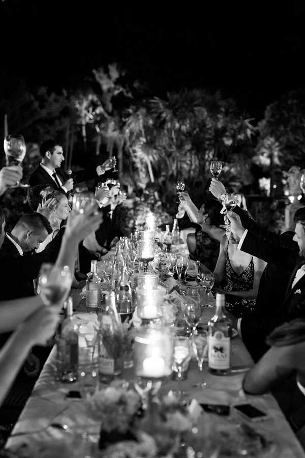 This black-and-white image captures an outdoor evening wedding reception toast, with approximately 20 or more guests seated along a long rectangular dining table raising their wine glasses simultaneously. The table is set with multiple wine glasses, candles providing the primary light source, wine bottles, and low floral centerpieces, though specific flower types are difficult to distinguish in the dark tones. The scene is set outdoors at night with tall tropical or palm-like trees visible in the dark background, lit from below. Guests are dressed in formal evening wear, with men in dark suits and women in dark dresses. The composition is a wide perspective shot taken from one end of the table looking down its length, creating a strong leading line, with dramatic contrast between the candlelit table and the very dark background.