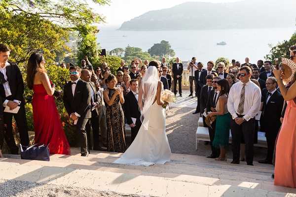 An outdoor wedding ceremony in progress, captured in a wide shot showing the bride walking down the aisle toward the groom and officiant. The ceremony is set on a terraced area overlooking a large body of water with hills visible in the background. The bride wears a fitted, sleek white gown with a long cathedral veil and carries a bouquet of cream and ivory blooms. Approximately 40-50 guests line both sides of a gravel aisle, dressed in formal attire including a bridesmaid or guest in a full-length red gown and another in a coral dress. White chairs are arranged on the right side of the aisle. The overall styling is classic and formal, with a warm, sunny Mediterranean atmosphere. Potential venue feature image.