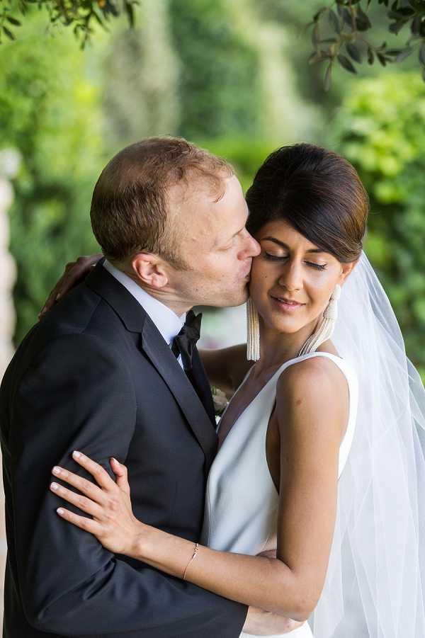 An outdoor couple portrait showing the groom kissing the bride on the cheek as she leans into him with her eyes closed. The groom wears a dark navy suit with a white dress shirt and dark tie, while the bride wears a sleeveless white dress with a simple V-neckline and a sheer white veil. Her dark hair is styled in a sleek updo and she wears long gold tassel drop earrings. The styling is modern and minimal. The shot is a close-up portrait framed against a blurred green foliage background.