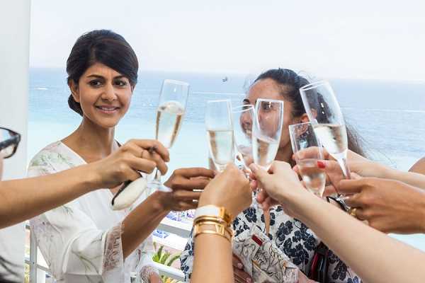 A group of women toast with champagne flutes during what appears to be a pre-wedding getting-ready celebration, likely a bridal party moment. The setting is an outdoor terrace or balcony with a sea view visible in the background. The woman on the left, likely the bride, wears a white floral robe and has her dark hair styled up, smiling directly at the camera. At least four to five women are visible, one wearing a black and white floral top. The shot is a close-up portrait-style image with the champagne glasses in the foreground, taken in bright natural daylight.