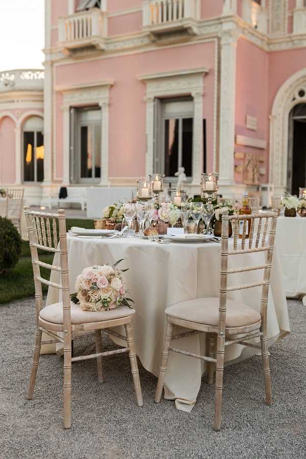 Outdoor wedding reception table setup photographed in a medium wide shot, positioned in front of a pink-facades villa with white ornamental detailing and arched windows. The round sweetheart or small guest table is dressed in a cream-ivory floor-length linen, surrounded by whitewashed chiavari chairs with champagne cushions. A bridal bouquet of blush and cream roses rests on one of the chairs. The centerpiece features low arrangements of blush roses, ivory blooms, and greenery alongside multiple gold mercury glass candleholders with lit pillar candles at varying heights. The table is set with crystal wine glasses, silver flatware, and white plates. The overall decor palette is blush, ivory, and gold, consistent with a classic, romantic styling approach. Potential venue feature image.