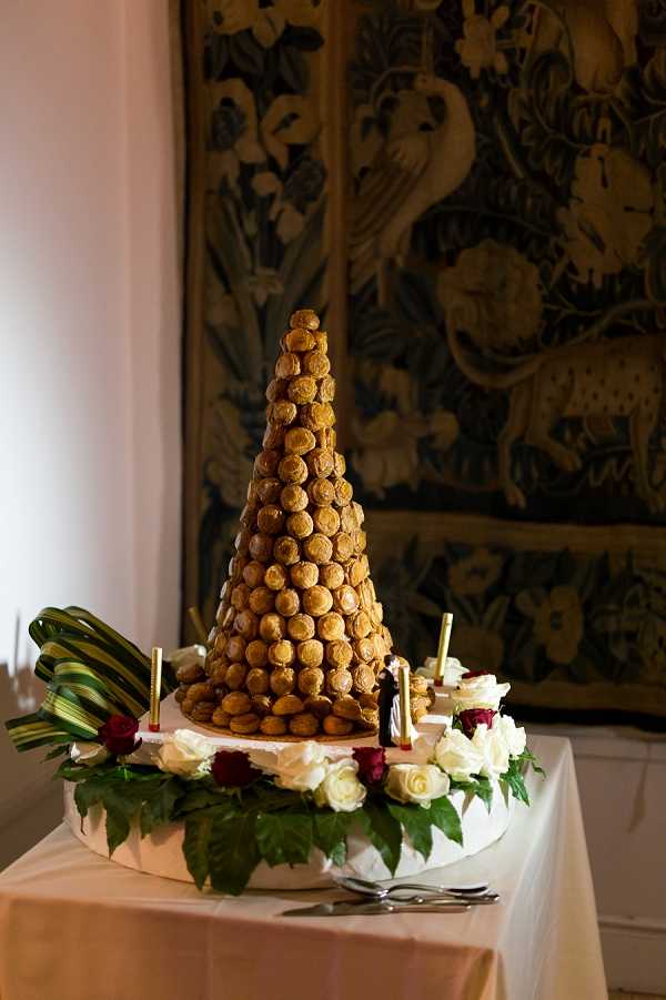 Close-up detail shot of a traditional French wedding croquembouche, a tall cone-shaped tower of caramel-glazed profiteroles displayed on a white cake board atop a table draped with a blush-pink linen. The base of the croquembouche is decorated with cream and deep red roses along with green foliage, and small gold taper candles are placed around it. A miniature bride-and-groom cake topper figurine is visible at the base of the tower. A large ornate tapestry featuring animals and floral motifs in muted gold, green, and brown tones hangs on the wall in the background, suggesting an indoor reception room in a historic venue such as a chateau.