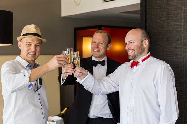 Three men toast with champagne flutes in what appears to be a hotel room or modern interior space during a getting-ready moment. The man on the left wears a white shirt and a straw panama hat with a black band, the man in the center is dressed in a black tuxedo with a black bow tie, and the man on the right wears a white dress shirt with a red bow tie and red suspenders. A champagne bottle is visible in the background on a surface. The shot is a medium portrait framing all three men from approximately the waist up, smiling toward the camera.