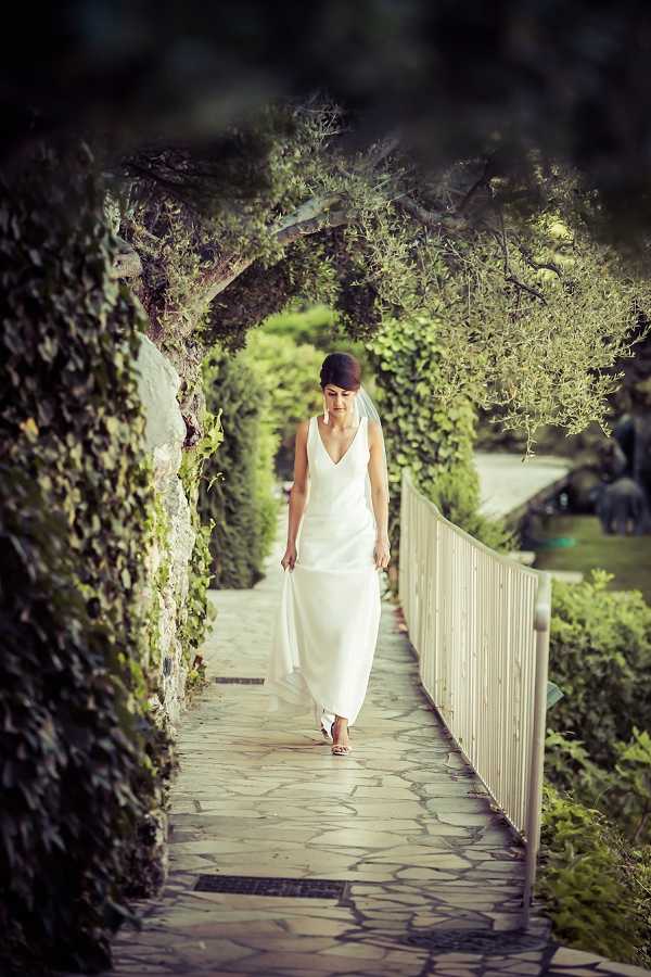 A bride walks alone along a narrow stone-paved garden path framed by a vine-covered archway, creating a natural tunnel effect around her. She wears a sleek, minimalist ivory sleeveless gown with a deep V-neckline and a flowing skirt, lightly holding the hem with one hand, paired with strappy sandals and drop earrings. Her dark hair is worn up and she looks downward as she walks. The composition is a full-length portrait shot taken from a distance, using the arched greenery to frame the subject and draw focus toward her.
