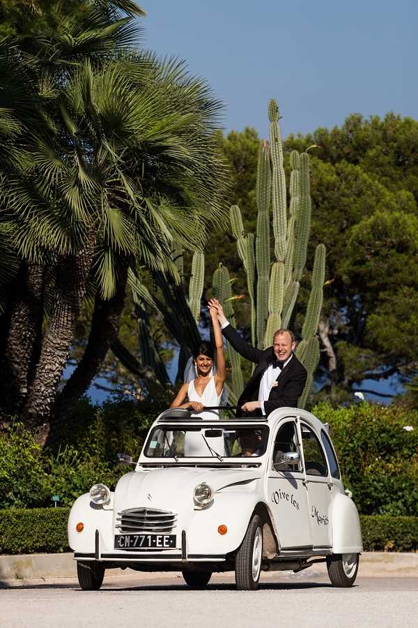 The bride and groom are standing up through the open roof of a vintage white Citroën 2CV, raising their joined hands in celebration. The car has 'Vive les Mariés' written on the door in script lettering and displays a French license plate. The bride wears a white halter-neck dress with a veil, and the groom is dressed in a black tuxedo with a white boutonnière. The couple appears to be on a driveway or road at an outdoor venue in the South of France, with tall columnar cacti and palm trees visible in the background, suggesting a Mediterranean coastal location. The shot is a full-length portrait framing the car and both figures, taken in bright daytime light.