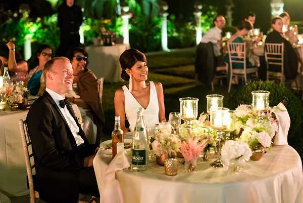 The bride and groom are seated at their sweetheart table during an outdoor evening reception, both laughing and visibly enjoying the moment. The bride wears a sleeveless white V-neck gown with her dark hair pulled back, and the groom is in a black tuxedo with a black bow tie. The table is decorated with low arrangements of blush pink and white flowers including what appear to be peonies and hydrangeas, accented with pink astilbe, set among gold mercury glass votive candle holders and pillar candles that cast warm light across the table. In the background, additional guests are seated at chiavari chairs around round tables, with white column structures illuminated by green uplighting visible among the surrounding garden. The shot is a medium close-up portrait capturing the couple from the side with the reception scene in soft focus behind them.