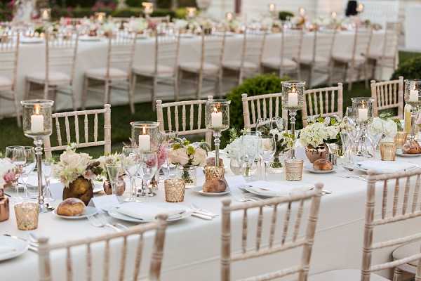 A wide shot of an outdoor wedding reception setup featuring multiple long banquet tables dressed in white linen, lined with natural wood chiavari chairs. The centerpieces combine white hydrangeas, blush and dusty pink florals, and greenery arranged in copper and gold vessels, alongside tall glass hurricane candle holders on silver candelabras, mercury glass votives, and small gold tealight holders. The overall decor palette is white, blush, champagne, and rose gold. Place settings include white plates, silver cutlery, and clear stemware. Several long tables extend into the background, all styled consistently, set in an outdoor garden or grounds setting with manicured hedging visible.