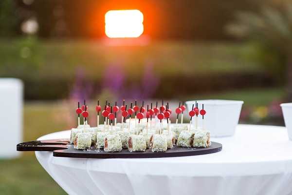 Close-up detail shot of a cocktail hour food display at an outdoor wedding reception. A round dark slate or wooden board holds rows of bite-sized canapés coated in what appears to be white sesame seeds or grated coconut, each topped with a decorative pick featuring a red ball finial and black stem. The board is placed on a high cocktail table draped with a white linen. The background is softly blurred with warm orange sunset light and hints of purple florals and white furniture visible in the garden setting.