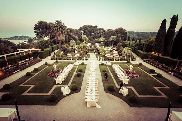 An aerial wide shot captures a formal outdoor wedding reception setup in a grand French garden at dusk. Long banquet tables dressed in white linens are arranged symmetrically along a central stone pathway flanked by geometric parterres, manicured hedges, and topiaries. The garden extends into a tree-lined perspective toward what appears to be a coastal waterline visible in the background, with warm ambient lighting beginning to illuminate the tables and surrounding landscape. The overall decor palette is white and cream, with touches of pink and red from planted flower beds bordering the lawn sections. Potential venue feature image.