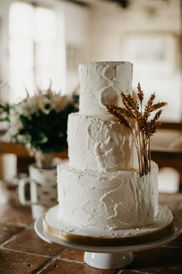 Close-up detail shot of a three-tiered wedding cake displayed on a white and gold cake stand, placed on a terracotta tile surface indoors. The cake is finished in textured white buttercream with an organic, rough-plastered appearance, and is topped with three stems of dried golden wheat. In the soft-focused background, a white ceramic mug holds a small arrangement of white blooms and greenery, alongside what appears to be a white pillar candle. The overall styling is rustic-organic with a natural, earthy palette combining white and warm gold tones.