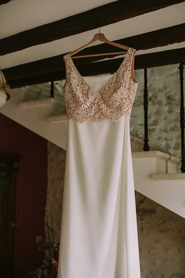 A getting-ready detail shot of a wedding dress hanging from a wooden hanger on a dark wooden ceiling beam inside what appears to be a rustic stone-walled venue. The gown features a deep V-neckline bodice in nude-toned floral lace with scalloped edges, transitioning into a sleek, column-cut ivory crepe skirt. The interior setting shows exposed dark timber beams, a wrought-iron staircase railing, and rough-cut stone walls, suggesting a historic French property. The composition is a centered portrait-style shot emphasizing the dress design.