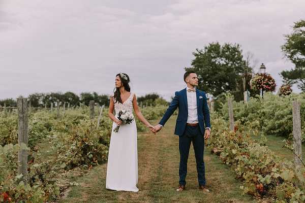 A couple portrait taken outdoors in a vineyard, with rows of grapevines extending on either side of a grass path. The bride wears a simple white V-neck gown with thin straps and a floral crown of small white and green blooms, holding a compact bouquet of white flowers and greenery. The groom wears a navy blue suit with a light pink dress shirt, a bow tie, and a pocket square, with a brown leather belt. The two stand hand-in-hand facing different directions, looking away from each other and from the camera. A decorative lamp post and hanging floral basket are visible in the background. The shot is a wide full-body portrait with a boho-relaxed styling aesthetic.