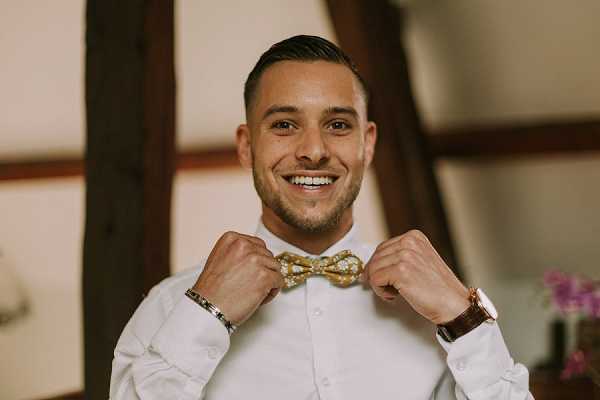 A groom is photographed indoors during the getting-ready portion of his wedding day, adjusting a gold and white patterned bow tie with both hands while smiling directly at the camera. He is wearing a white dress shirt, a gold/rose-tone watch on his right wrist, and a silver bracelet on his left wrist. The background suggests a rustic interior with dark wooden beams visible, and a small arrangement of purple flowers is partially visible in the right background. This is a close-up portrait shot with a shallow depth of field keeping the groom in sharp focus.