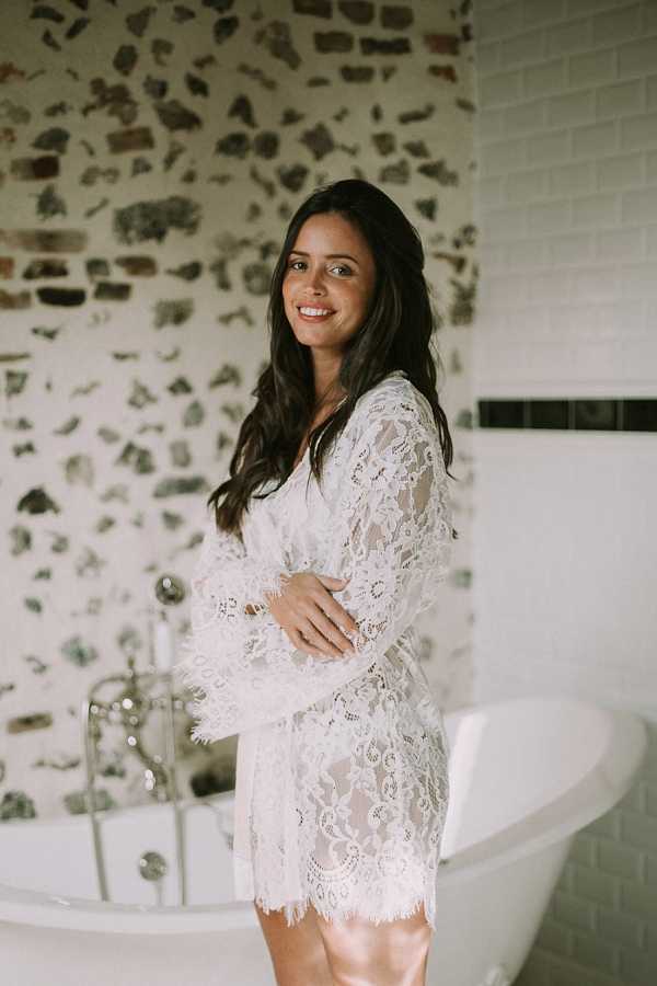 A bride is photographed during her getting-ready preparations, standing in a bathroom in front of a freestanding white clawfoot bathtub with silver fixtures. She is wearing a white lace robe with long sleeves and scalloped hem detailing, with her dark hair worn loosely down. The bathroom features an exposed stone wall alongside white subway tiles with a dark tile accent border, giving a rustic-meets-classic feel to the space. The shot is a medium portrait framing her from mid-thigh up, with natural light illuminating the scene.