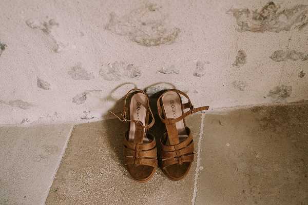 A close-up detail shot of a pair of tan brown leather strappy sandals with a low wedge sole, placed on a stone floor against a textured white plaster wall. The sandals feature multiple cross straps and ankle buckle closures, styled in a bohemian or rustic aesthetic. The shot is a flat-lay-style composition taken from slightly above, with natural ambient lighting highlighting the warm tones of the leather against the aged stone surface.