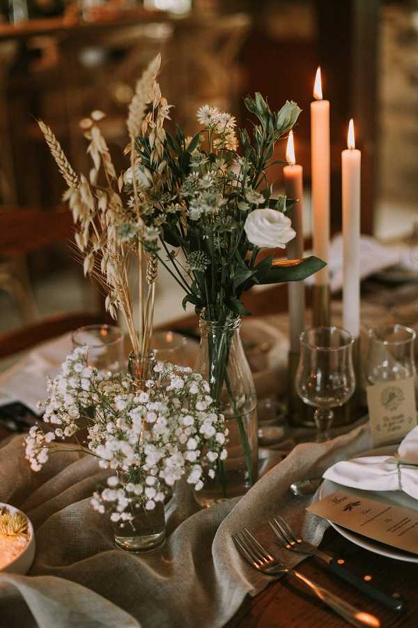 Close-up detail shot of a wedding reception table centerpiece with a rustic, boho styling theme. The centerpiece features multiple glass bottles and vases holding an arrangement of dried wheat stalks and pampas-style grasses, white roses, white lisianthus buds, small blue thistle flowers, yellow wildflowers, and a separate small jar of white baby's breath. Three tall cream taper candles are lit and placed in candlestick holders alongside the florals, casting warm amber light across the setup. The table is dressed with a natural linen or burlap runner in a muted sage-grey tone over a dark wood surface, and place settings include dark slate-toned plates, a printed kraft paper menu card, white linen napkins, and silver cutlery.
