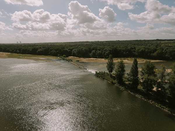 Aerial wide shot of a wide river with a forested riverbank and sandbanks visible in the background. No people, wedding decor, or wedding-related activity are visible in this image. The shot appears to be a landscape/environmental photograph, likely taken by drone to establish the surrounding geography near a venue. No intentional wedding styling elements are present.