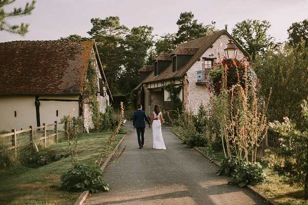 A bride and groom walk hand-in-hand away from the camera along a paved pathway leading toward a cluster of rustic stone and half-timbered farm buildings, likely a domaine or ferme venue in the French countryside. The bride wears a white gown with an open or low back, and the groom is dressed in a navy suit. The pathway is flanked by tall flowering plants and lush greenery, with a vintage-style lantern post visible near the main building. The shot is a wide, candid portrait taken at dusk with a warm, soft light. Potential venue feature image.