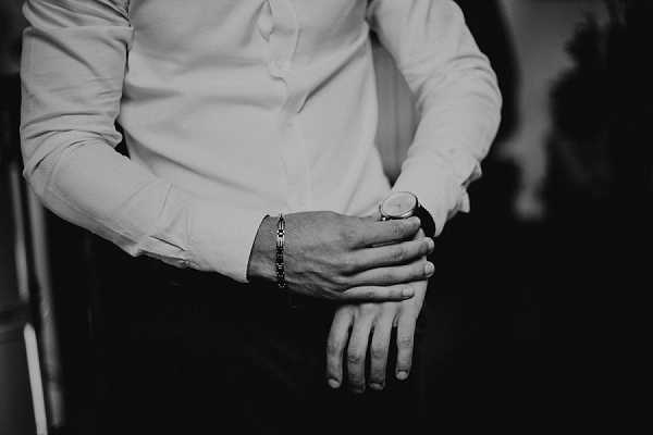 A black-and-white close-up detail shot of a groom getting ready, captured from the torso down. He is adjusting his watch cufflink area while wearing a crisp dress shirt and dark trousers, with a chain bracelet visible on his wrist. The image features high contrast with deep shadows in the background, drawing focus to the hands and accessories. The composition is a tight detail/close-up portrait emphasizing the groom's preparation ritual.