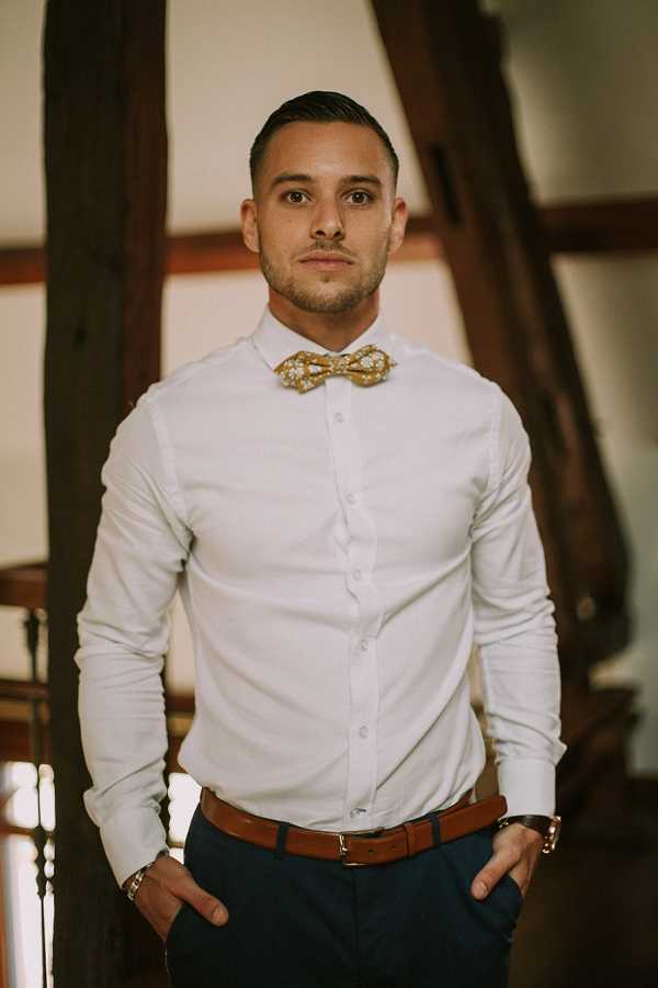 Portrait of a groom during the getting-ready phase, standing indoors with his hands in his pockets and looking directly at the camera. He is wearing a white fitted dress shirt, navy trousers, a tan leather belt, and a gold patterned bow tie with a small floral or dotted print. A watch and bracelet are visible on his wrists. The background features exposed dark wood timber-frame beams, suggesting a barn or rustic venue interior. The shot is a mid-length portrait with a shallow depth of field that keeps the background softly out of focus.
