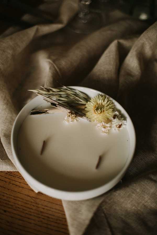 Close-up detail shot of a large white ceramic multi-wick candle decorated with dried botanicals on its surface, including dried wheat stalks, a pale yellow strawflower, and small white dried daisy-like blooms. The candle sits on a wooden slice and is surrounded by a loosely draped taupe linen cloth, with the base of a glass vessel partially visible in the background. The overall decor palette is earthy and muted — cream, taupe, and dried natural tones — consistent with a rustic or boho styling theme. A stemmed glass is partially visible in the upper background, suggesting a table setting context.
