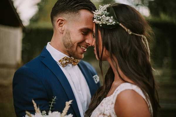 A close-up couples portrait of a bride and groom touching foreheads and smiling, shot outdoors with a softly blurred background. The groom wears a navy blue suit with a white dress shirt and a gold floral-patterned bow tie, with a matching pocket square. The bride wears a lace-detailed white dress and a floral crown of white baby's breath, with long dark hair worn down; she holds a boho-style bouquet featuring dried pampas grass and greenery. The styling has a boho-romantic aesthetic with warm, golden-toned natural light. Medium close-up portrait framing cuts off just below the shoulders.
