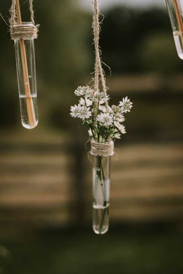 Close-up detail shot of rustic wedding decor featuring glass test tubes suspended by natural jute twine. The central tube holds small white wildflowers, likely astrantia, with delicate star-shaped blooms, while the tube to the left contains water and thin wooden sticks or skewers. The twine is wrapped around the neck of each tube to create the hanging mechanism. The background is blurred and outdoor, consistent with a garden or rural setting. The overall styling is rustic and bohemian, using raw, natural materials.
