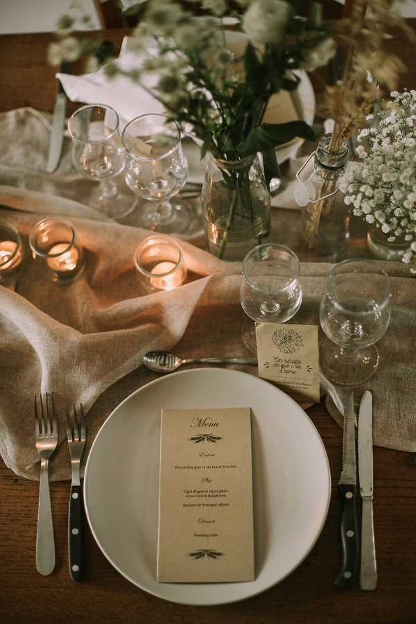 Close-up detail shot of a wedding reception place setting on a dark wood table. A white ceramic plate holds a kraft paper menu card printed in French, listing courses including foie gras, agneau, fromages, and Wedding Cake. The table is styled with a loosely draped natural linen runner in a warm sand tone, and small votive candles in amber glass holders provide warm candlelight. Floral centerpieces include white gypsophila (baby's breath), green eucalyptus, and dried pampas-style stems in small glass bud vases and bottles. Two wine glasses and a water glass are placed at the setting alongside a small yellow place card. Cutlery includes two forks on the left and a knife and spoon on the right. The overall decor palette is natural and rustic — warm neutrals, kraft paper stationery, linen, and candlelight.