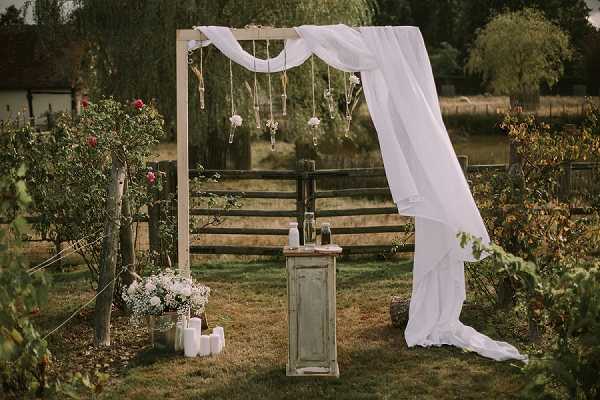 An outdoor ceremony setup in a garden setting, showing a wooden arch draped with flowing white fabric that extends to the ground on one side. The arch is decorated with hanging glass bud vases containing small blooms, suspended by twine or ribbon. A rustic distressed grey-green wooden cabinet serves as the ceremony altar, holding bottles and small decorative items on top. To the left of the arch, a white arrangement of baby's breath and mixed white flowers sits in a pot on the grass, surrounded by several white pillar candles of varying heights. The overall styling is boho-rustic with a white and natural wood palette. Wide shot capturing the full ceremony installation with a wooden fence and garden foliage visible in the background.