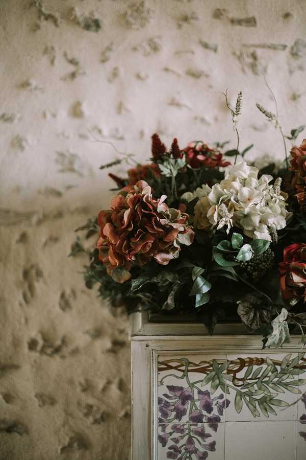 A close-up detail shot of a floral arrangement sitting atop a decorative painted piece of furniture, likely a mantelpiece or cabinet featuring hand-painted wisteria motifs in purple and green on a cream background. The arrangement includes rust-toned and cream hydrangeas, deep red celosia or amaranth, green foliage, and small berry-like accents, creating an autumnal, rich color palette. The background is a textured, aged plaster wall with a mottled appearance. The overall styling suggests a rustic or vintage aesthetic with an earthy, warm tone.