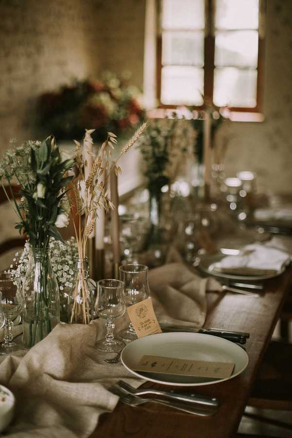 A close-up detail shot of a rustic indoor wedding reception table setting. The table is styled with a natural linen runner, clear glass bud vases holding dried wheat stems, baby's breath, and green foliage, alongside tall tapered blush-toned candles in brass candlesticks. Each place setting features a white ceramic plate with a printed menu card, kraft paper name cards, dark-handled cutlery, and clear stemmed glassware. In the background, a larger arrangement of deep red and green florals is visible near a wooden-framed window, contributing to an earthy, rustic-botanical decor palette with warm amber tones throughout.