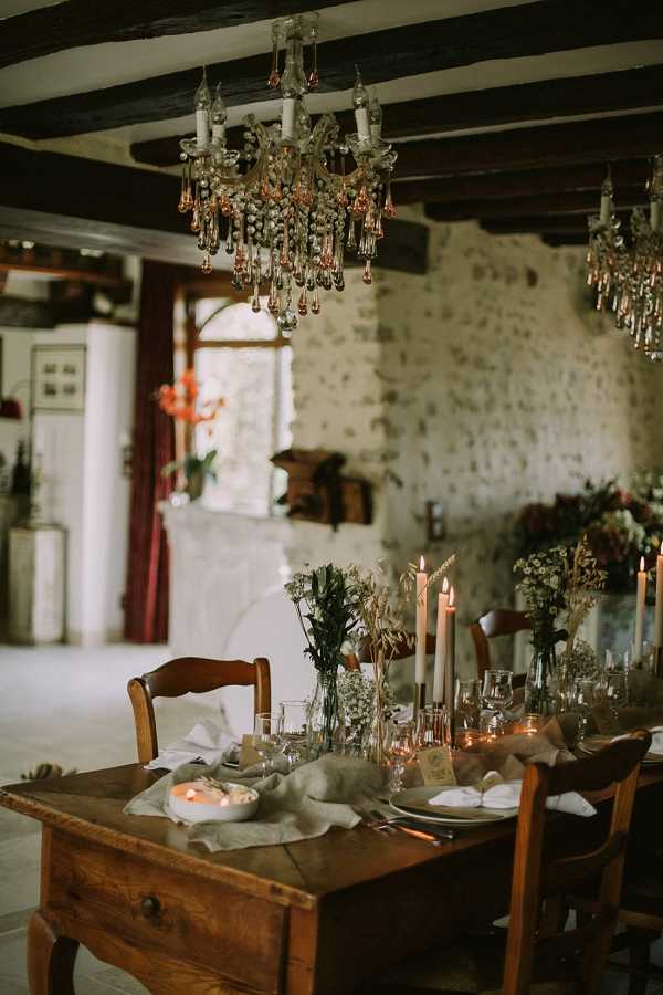 A reception tablescape detail shot taken indoors in a rustic French venue with exposed stone walls and dark wooden ceiling beams. A long antique wooden farmhouse table is set with a linen runner in a natural oat tone, stoneware plates, linen napkins, and an assortment of glassware. The centerpiece features clusters of bud vases holding wildflowers, baby's breath, wheat stems, and greenery, alongside tall ivory taper candles in varying heights, with several already lit and casting warm candlelight. Two vintage crystal chandeliers with amber and rose-tinted droplets hang above the table, adding to the warm, ambient lighting. The overall decor palette is earthy and muted — natural linens, warm wood, dried and wild botanicals — giving the setting a rustic, organic aesthetic. Red curtains and a glimpse of florals in deep red tones are visible in the background through a doorway.