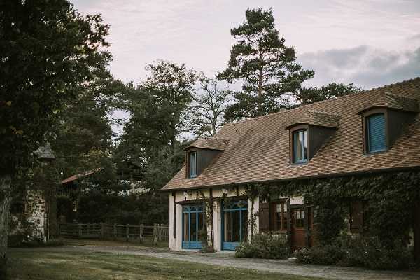 Wide exterior shot of a French country property featuring a low-rise building with a steep brown tile roof, dormer windows, and blue-painted window and door frames. The facade is partially covered in climbing ivy, and the building sits along a gravel courtyard. No people are visible in the frame. The image has a muted, slightly overcast tone with a moody atmosphere typical of film-style editing. Potential venue feature image.