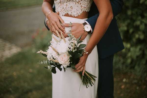 A close-up portrait of a couple embracing outdoors, with the groom's arms wrapped around the bride from behind. The bride is wearing a two-piece gown with a lace crop top and a white skirt, paired with a rose-gold watch and bracelets, and holds a loose, garden-style bouquet featuring blush roses, white protea, pampas grass, astilbe, and eucalyptus foliage. The groom is dressed in a navy blue suit. The composition is a mid-shot focused on the couple's torsos and the bouquet, with a softly blurred green outdoor background.