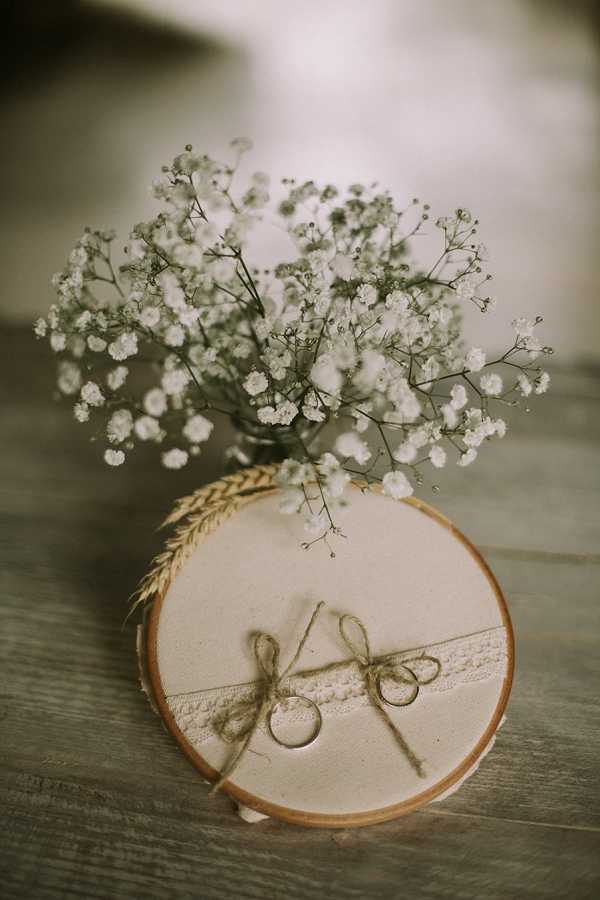 A close-up detail shot of a rustic wedding ring display. Two wedding bands are tied with twine bows onto a circular wood slice decorated with a strip of cream lace trim, resting on a weathered wooden surface. A small bunch of white gypsophila (baby's breath) in a glass vessel and a dried wheat stem are arranged behind the ring bearer. The styling is rustic and natural, using raw materials — wood, jute twine, lace, and dried botanicals — in a warm, neutral palette.