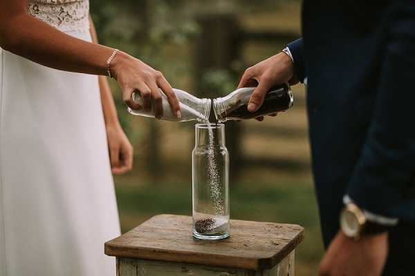 A close-up detail shot of a unity sand ceremony during an outdoor wedding. The bride, wearing a white dress and a delicate bracelet, and the groom, in a navy suit with a watch visible, are each pouring sand from small vessels — one white, one dark — into a clear glass bottle partially filled with reddish-brown and white sand. The bottle sits on a weathered wooden pedestal. The background is softly blurred greenery, suggesting a garden or wooded outdoor ceremony setting.
