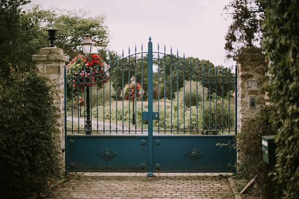 Wide shot of a decorative wrought-iron entrance gate painted in teal/dark blue-green, set between stone pillars on a cobblestone driveway. A black lamp post to the left of the gate holds a hanging basket of red and pink flowers. The gate features ornate scrollwork panels at the base and arched ironwork with spear-top finials across the top. Beyond the gate, a landscaped garden with dense greenery and flowering shrubs is visible. No people are present in the frame. Potential venue feature image.