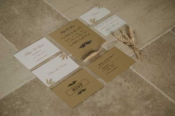 A flat lay detail shot of a wedding stationery suite arranged on a stone tile floor. The collection includes a main invitation, save the date card, RSVP card, dinner menu, and additional insert cards, all featuring a kraft brown and white color palette with gold and black wheat motif illustrations. A small bundle of dried wheat stalks tied with sheer ribbon is placed alongside the suite as a styling prop, reinforcing the rustic, countryside theme. The overall design aesthetic is rustic and nature-inspired, consistent with a French rural or farmhouse wedding style.