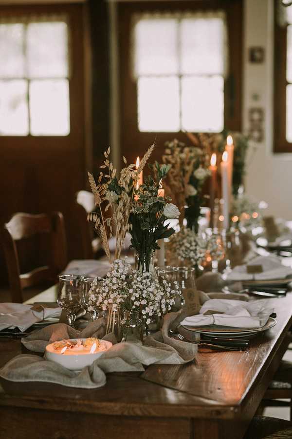 A close-up detail shot of a rustic wedding reception tablescape set on a dark wood farm table in an indoor venue with wood-framed windows visible in the background. The centerpiece arrangement features clusters of white baby's breath, dried wheat stalks, greenery, and a single white rose displayed in glass bud vases of varying heights, styled in a relaxed, boho-rustic aesthetic. Tall terracotta-toned taper candles are lit along the table runner, which is made of loosely draped linen in a muted grey-green tone. Place settings include dark charger plates layered with white linen napkins, wine glasses, and black flatware, with a small white bowl containing an orange-toned appetizer visible in the foreground.