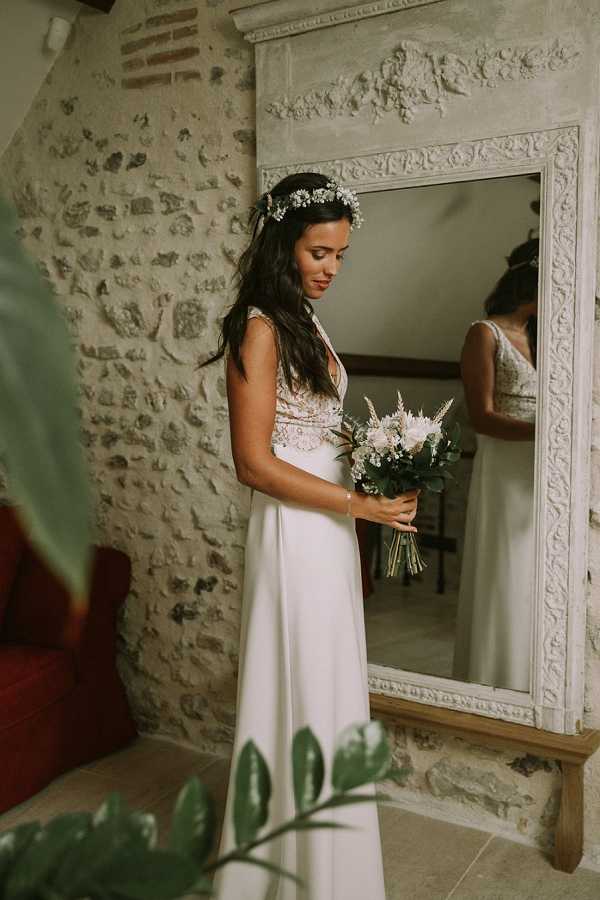 A bride stands indoors in front of a large ornate white-painted mirror, looking down at her bouquet during what appears to be a getting-ready moment. She wears a floor-length white gown with a lace or embroidered bodice and a flowing skirt, paired with a floral crown of small white daisy-like flowers and greenery in her long dark hair. Her bouquet is a loosely arranged mix of blush and white blooms, feathery white astilbe or similar textural flowers, and eucalyptus greenery. The setting features exposed stone walls and a decorative carved white fireplace surround, giving the space a rustic yet refined feel. This is a three-quarter portrait shot with the mirror reflection visible behind her, adding depth to the composition.