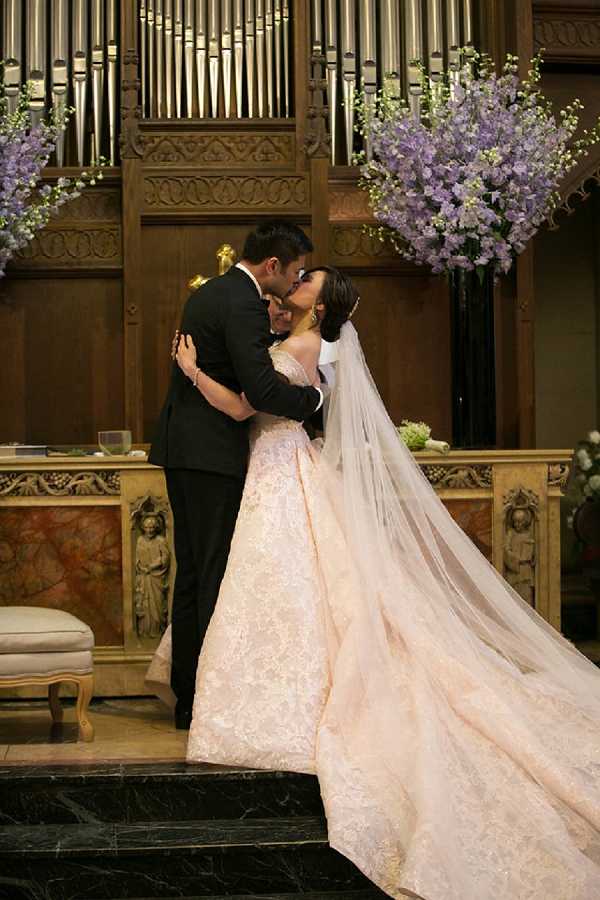 The bride and groom share their first kiss during an indoor church ceremony, standing at the altar steps in front of a large pipe organ with ornate carved wooden paneling. The bride wears a blush pink lace ball gown with an off-the-shoulder neckline, a long cathedral-length veil, and a jeweled headpiece, while the groom is dressed in a classic black tuxedo. Large arrangements of lavender and light purple delphinium-style flowers on tall black stands flank the altar on both sides, adding height and a soft purple color palette to the ceremony decor. The altar features gilded carved stonework with relief figures, and a cream upholstered stool is visible to the left. The shot is a full-length portrait taken from a slight distance, capturing the full volume of the bride's gown and veil.